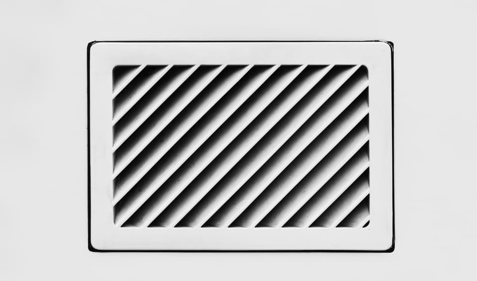 A white air vent grille with angled louvers set into a white wall
