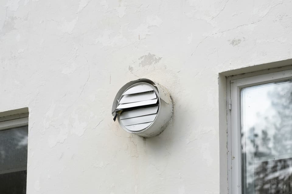 A round louvered exhaust vent on a white stucco house wall, one louver bent loose